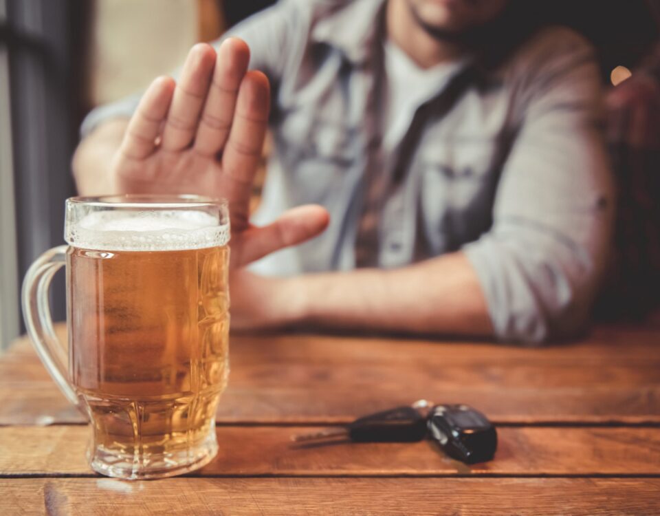 Das Bild zeigt einen Mann, der an einem Tisch sitzt und eine ablehnende Geste Richtung eines vollen Bierglases macht. Vor ihm liegt ein Autoschlüssel.